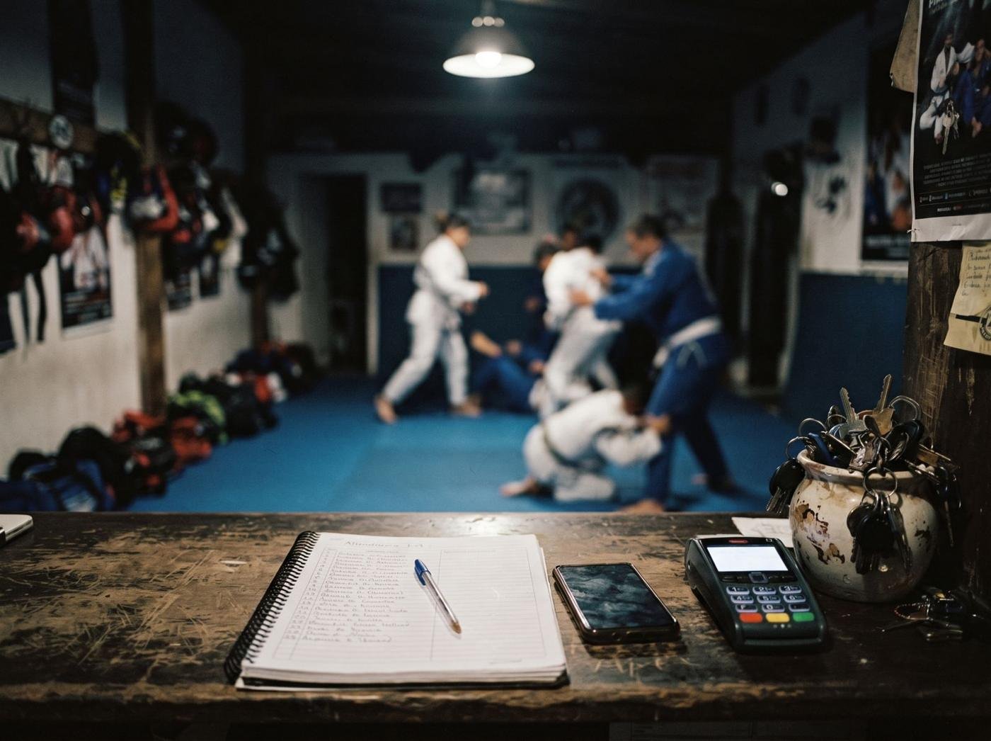 Caderno de chamada e maquininha no balcão da recepção, com uma aula de jiu-jitsu ao fundo